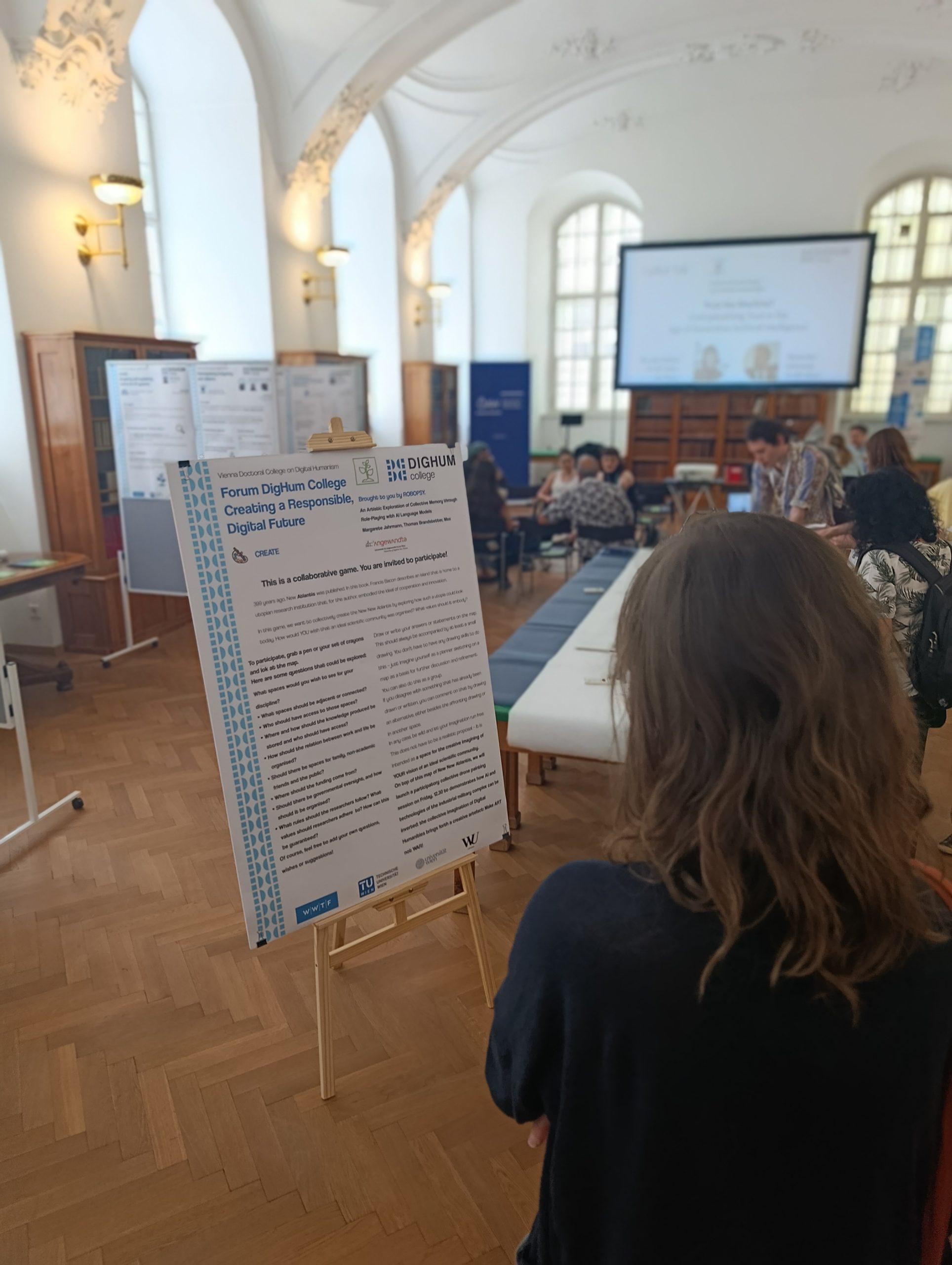 Person with shoulder-length hair standing in a room with arched windows and wooden floor looking at a poster on an easel titled 'Forum Digihum College Creating a Responsible Digital Future'